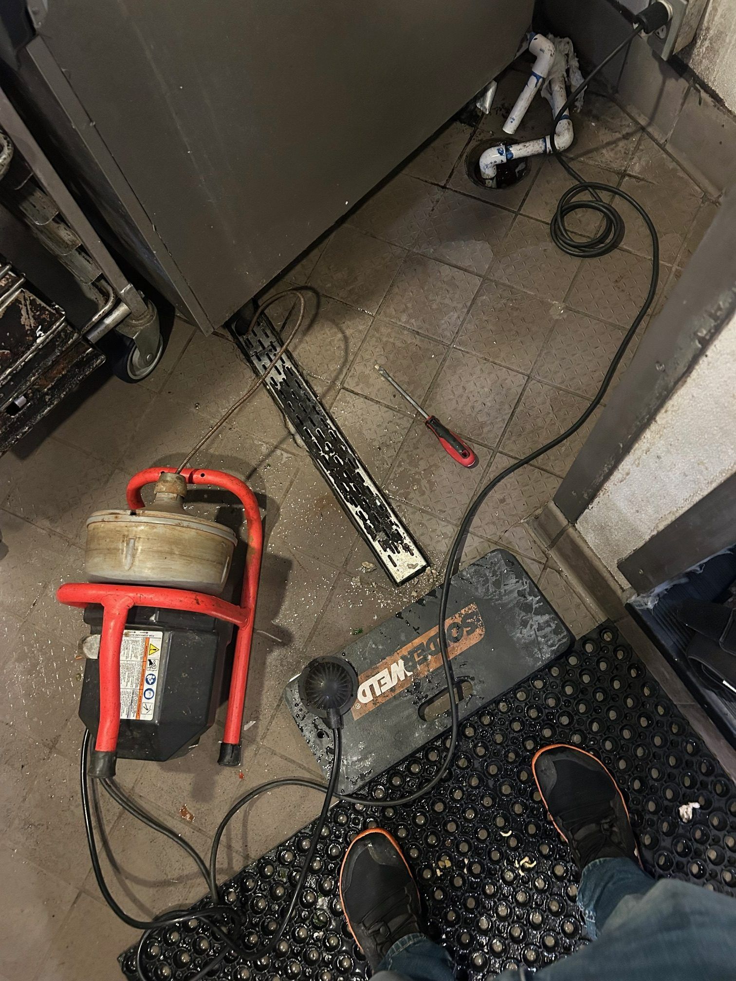 A drain cleaning machine on a floor with debris, next to a metal grate and a person's feet.