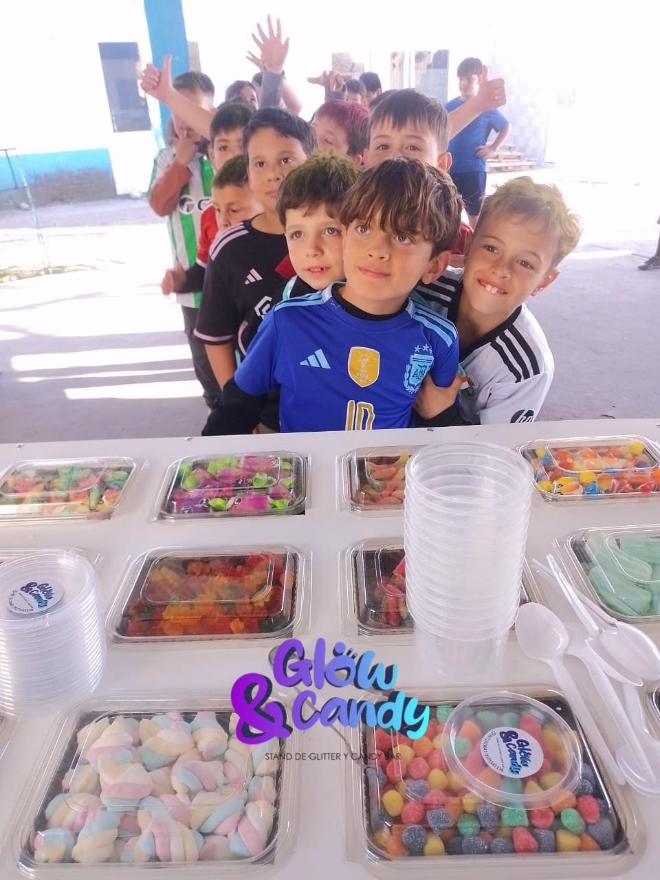 Niños en una barra de dulces, sonriendo, mirando a la cámara. Caramelos de colores en bandejas sobre una mesa.