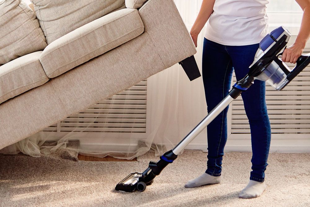 Young Woman Cleaning Carpet Under Sofa With Vacuum Cleaning — Big Red Carpet & Property Maintenance In Bowen, QLD