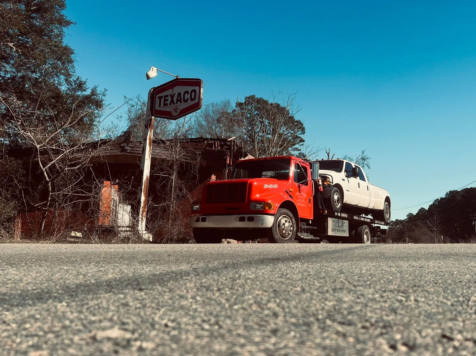A red flatbed tow truck carrying a white pickup truck parked on a roadside in front of an old, weathered Texaco sign.