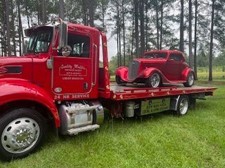 Red tow truck transporting a matching red classic car on a grassy area.