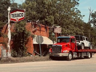 Red tow truck carrying a white Jeep in front of an old, abandoned Texaco gas station.