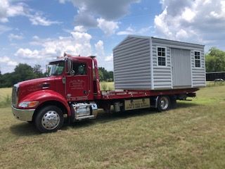 A bright red flatbed truck parked in a grassy field, carrying a light gray portable shed with two windows and a door.
