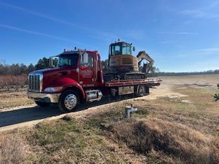 A red tow truck carrying a small yellow excavator on its flatbed, parked on a dirt path in a rural, sunny field.