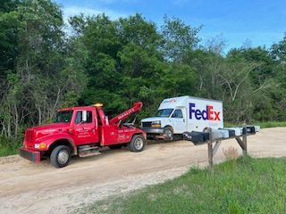 Red tow truck towing a white FedEx truck on a dirt road, near green trees and mailboxes.