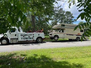 A white tow truck towing an RV on a road, both vehicles are in daylight, green grass and trees in the background.