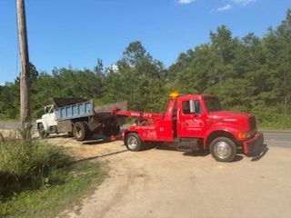 Red tow truck towing a white dump truck on a dirt road, trees and blue sky background.