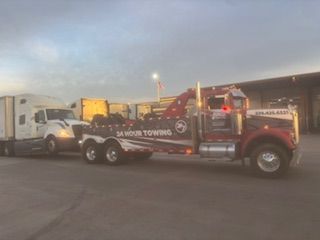A red heavy-duty tow truck is pulling a white semi-truck in a parking lot under a sunset sky.