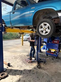 Mechanic under a blue pickup truck on a lift, working on the vehicle in a garage.