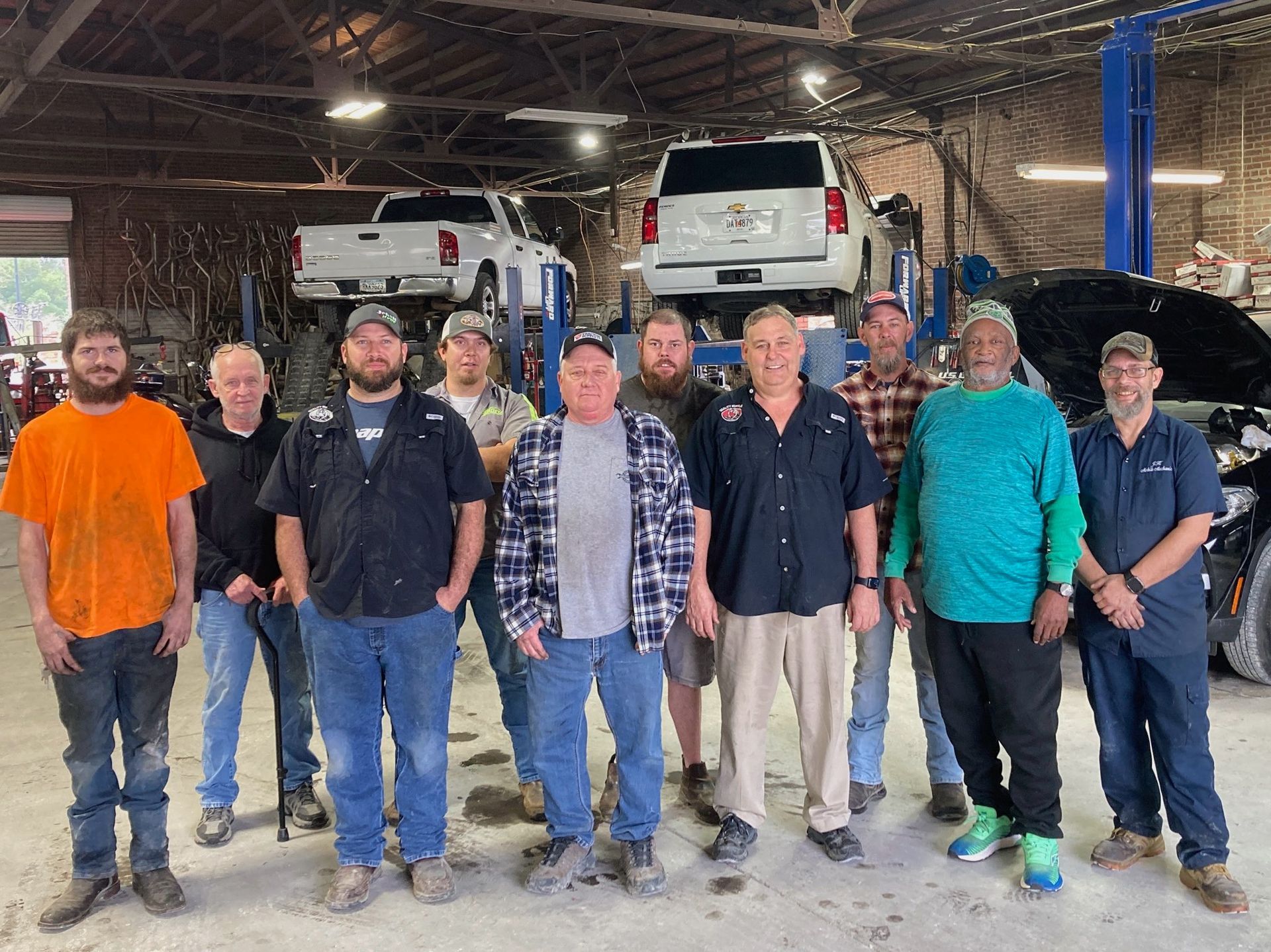A group of people wearing casual work clothes stand together in an auto repair shop with vehicles lifted on racks.