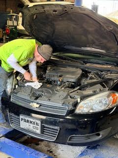 Mechanic in green shirt examines car engine bay, car parked inside a shop.