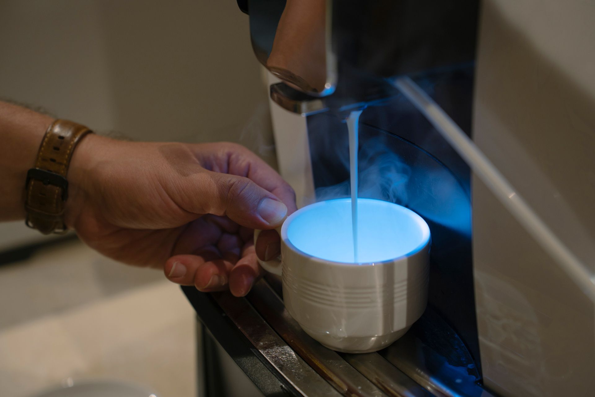 Hand holding a cup under a machine dispensing a white liquid, with a blue light inside the cup.