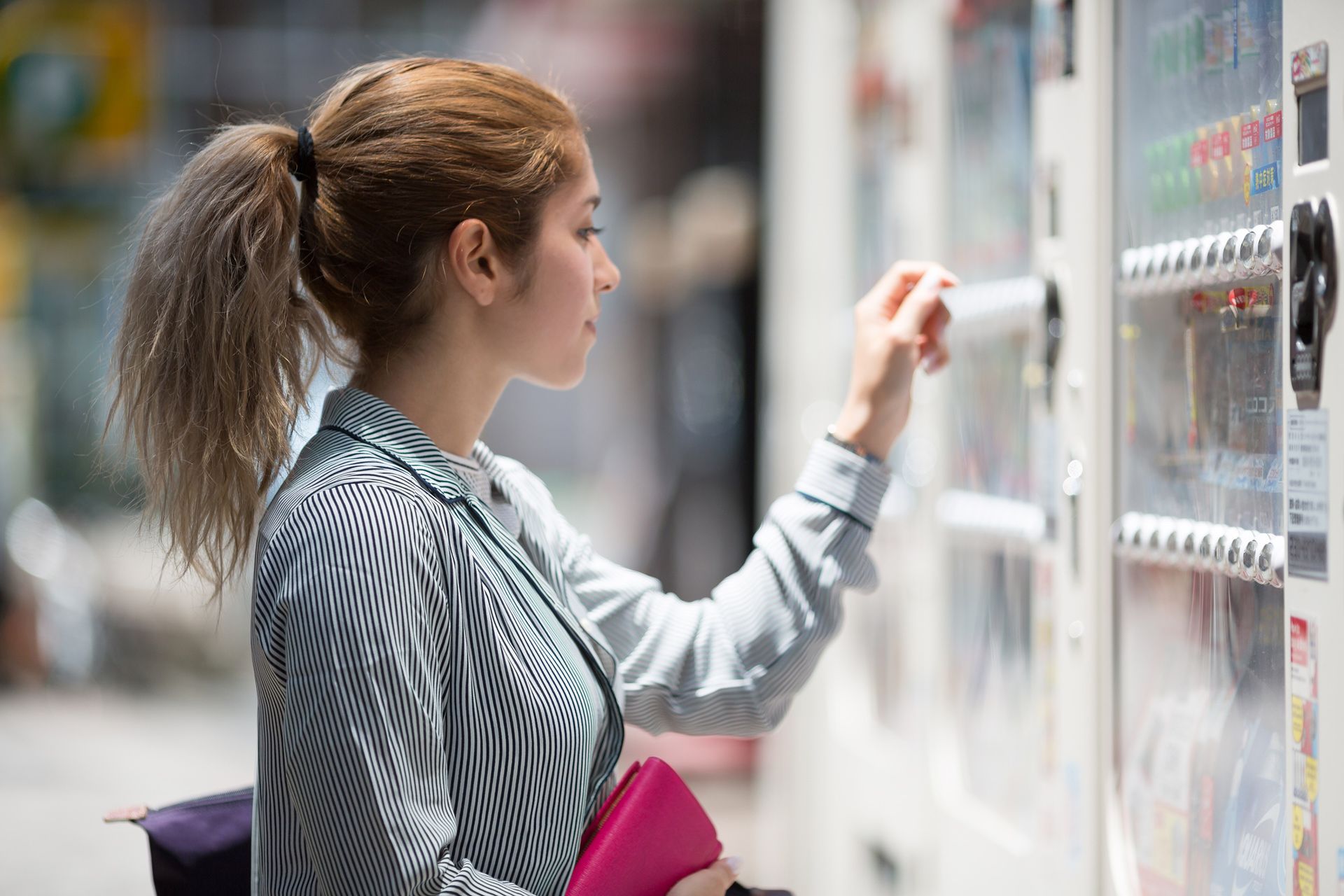 Woman selecting a drink from a vending machine, wearing a patterned shirt and with her hair in a ponytail.