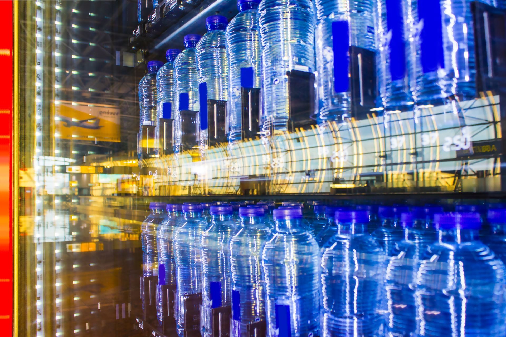 Rows of bottled water bottles reflect an airport terminal with yellow and blue hues.