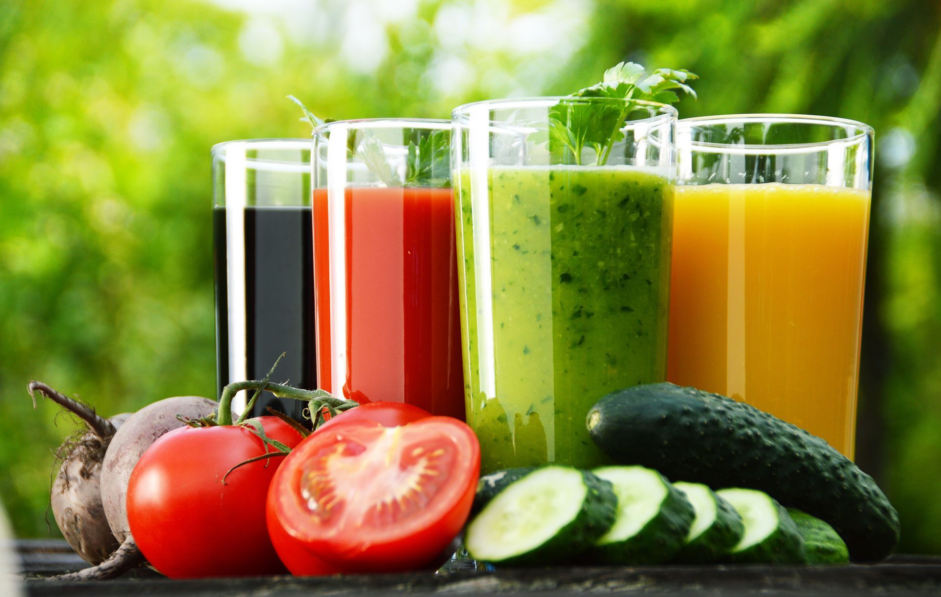 Glasses of colorful juice next to vegetables: beet, tomato, cucumber. Green outdoor setting.