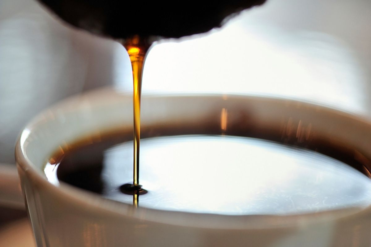 Dark liquid pouring from a dark object into a white mug filled with the same liquid.