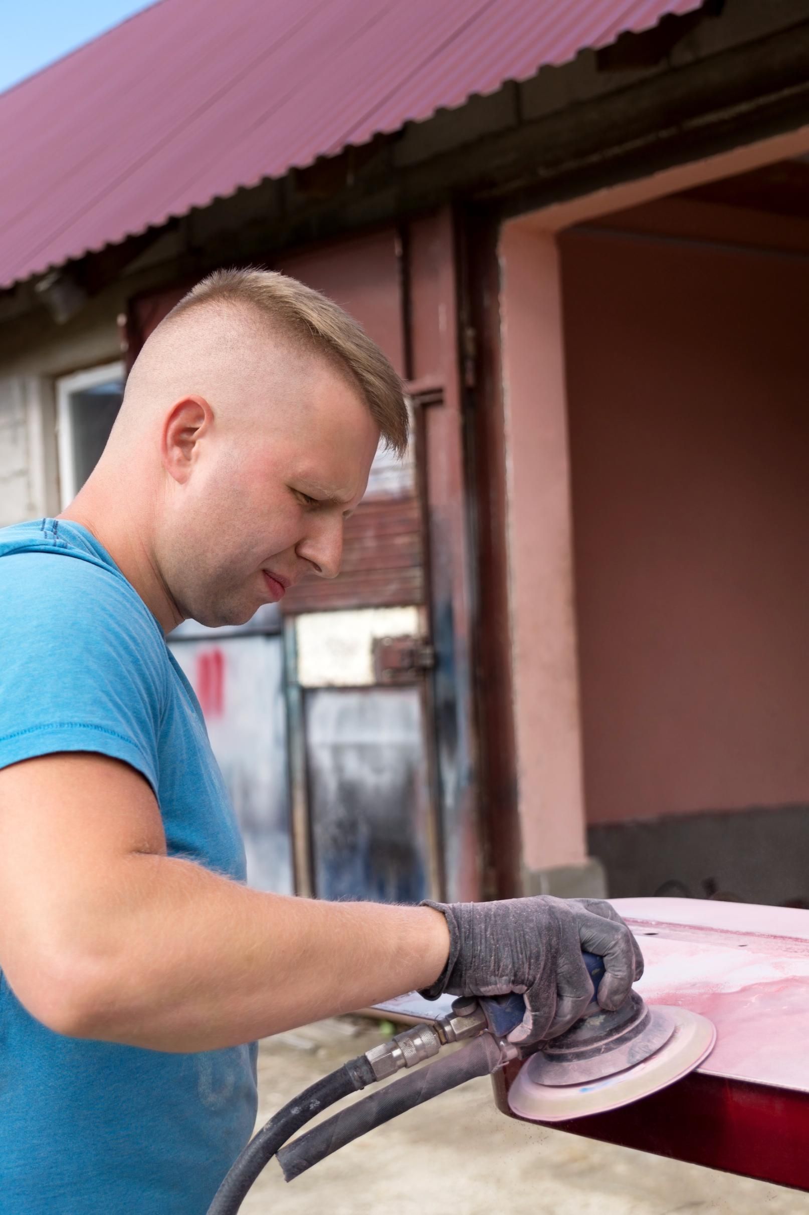 A man is sanding a red car with a sander.
