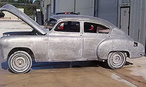 An old silver car with the hood up is parked in a parking lot.