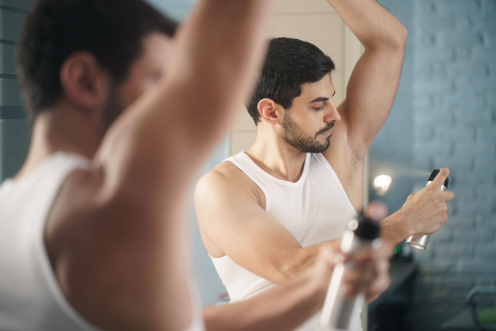 A man is spraying deodorant on his armpit in front of a mirror.