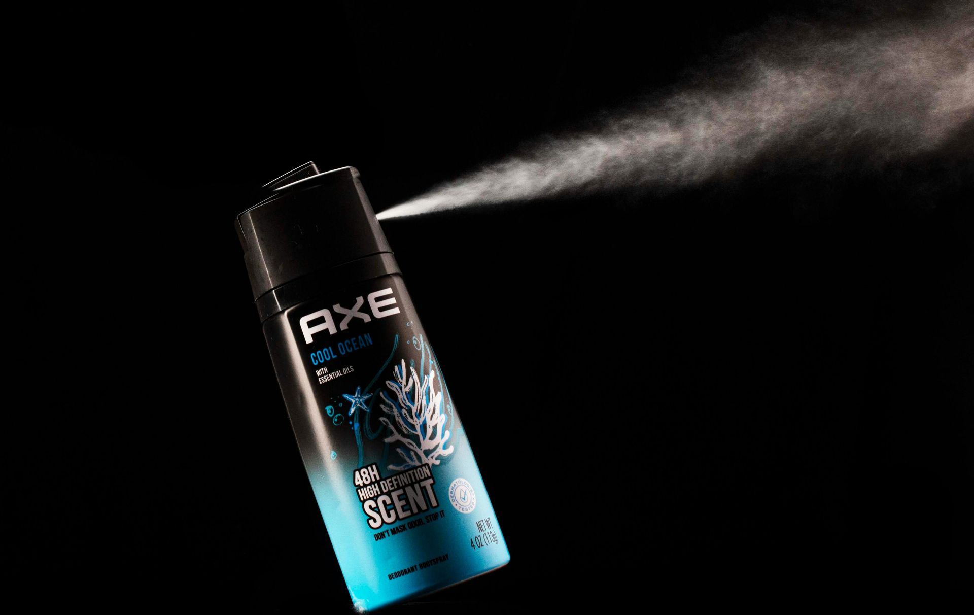A black and teal Axe body spray aerosol can angled to the left, spraying a mist into a dark background.