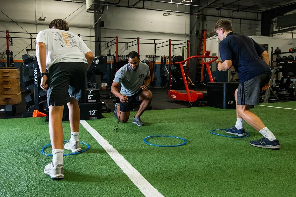 Athletes doing agility drills on indoor turf with cones and sleds in a gym.