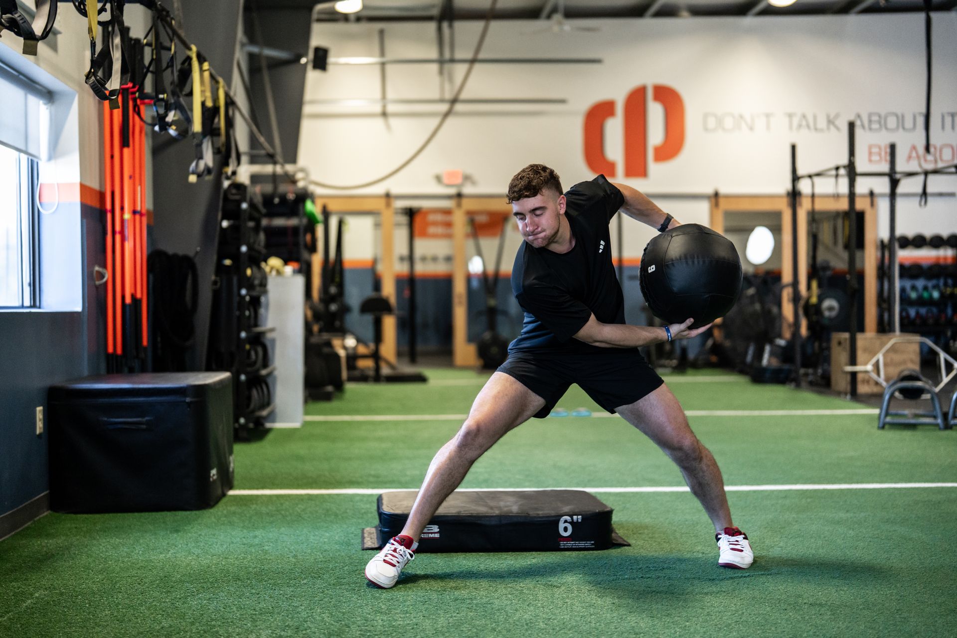 Man doing a kettlebell exercise in a gym, standing on turf with workout equipment around him.