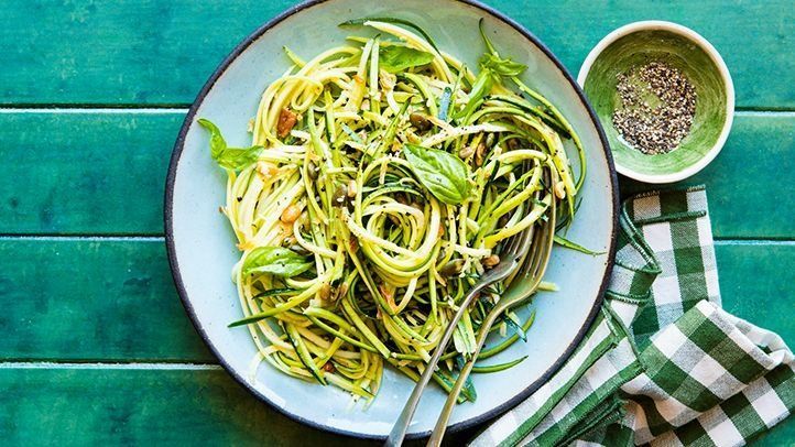 A bowl of zucchini noodles with herbs and spices, set on a green wooden table with a checkered napkin and small bowl.