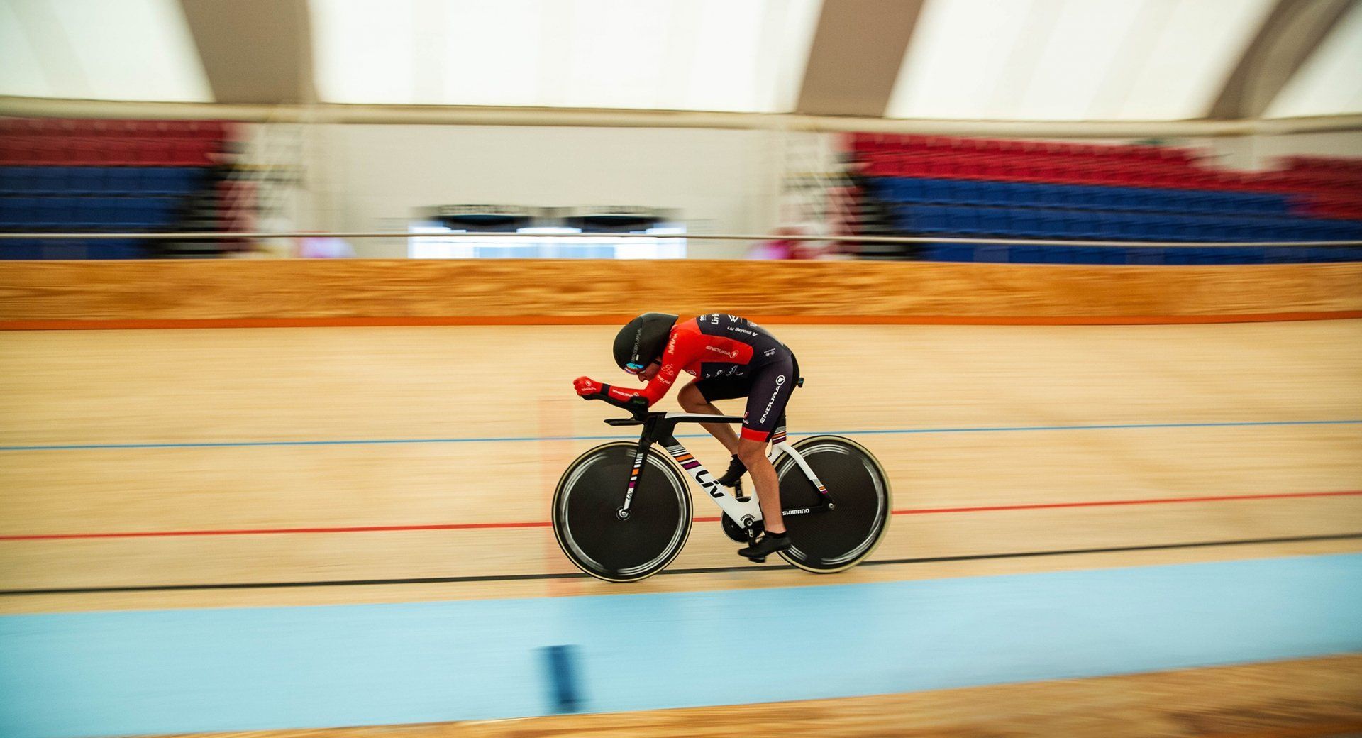 Cyclist in red and black racing on a velodrome track, leaning low over the handlebars.