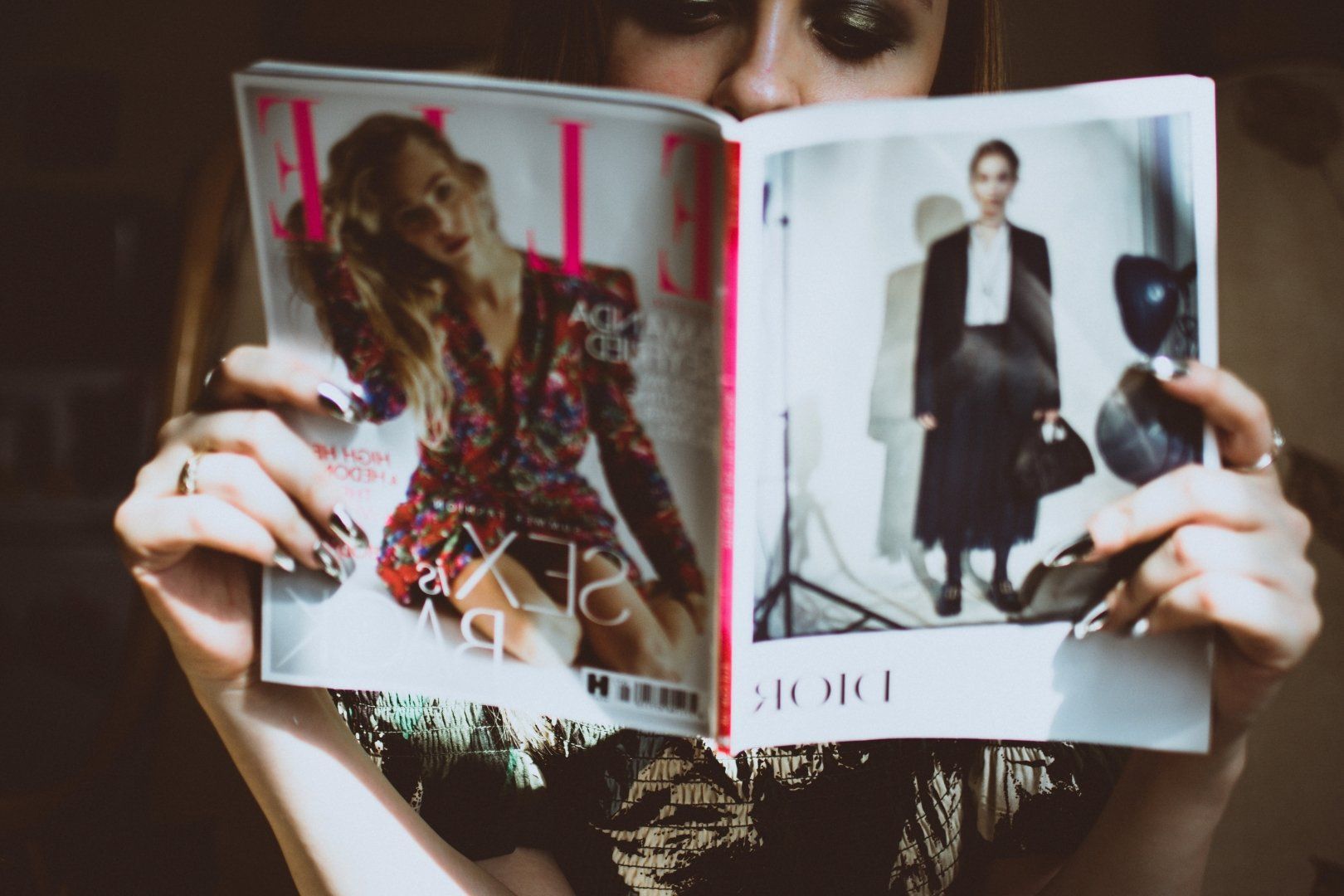 Person holding open fashion magazine in front of face, showing two model photos on the pages.