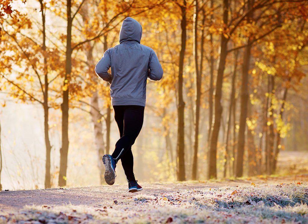 Person jogging on a leaf-covered forest path in a gray hoodie, surrounded by autumn trees with golden leaves