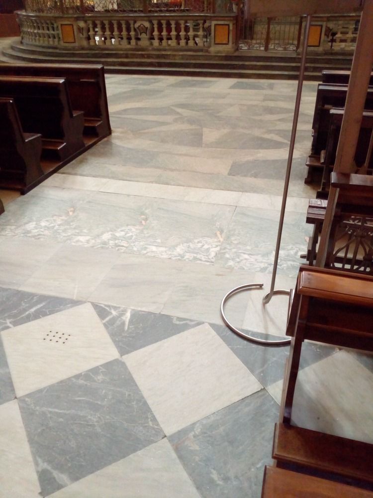 Vista interna del pavimento di una chiesa con piastrelle di marmo, banchi e un supporto di metallo.