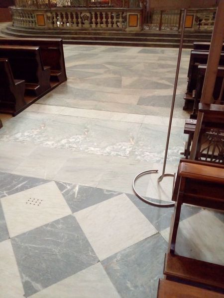 Vista interna del pavimento di una chiesa con piastrelle di marmo, banchi e un supporto di metallo.