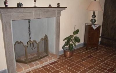 Camino con focolare in mattoni, paravento in metallo e mobiletto in legno con lampada, su pavimento in piastrelle di mattoni.