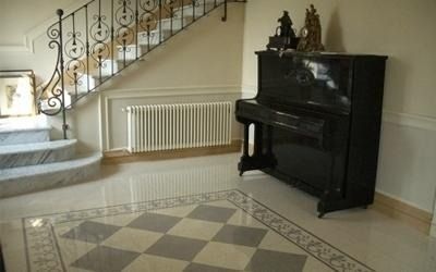 Un pianoforte nero in un atrio con pavimento in marmo a scacchiera, scale e un termosifone.