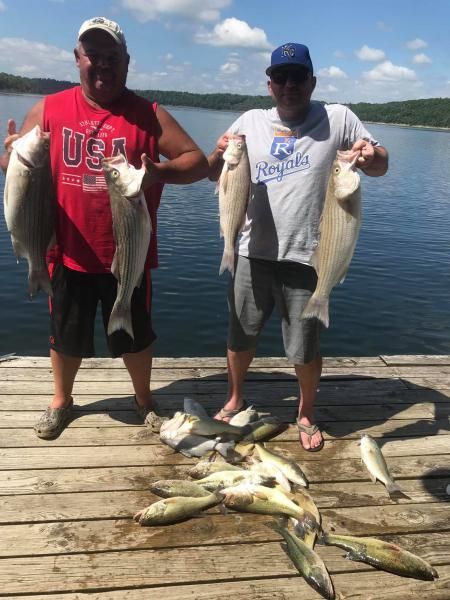 Two anglers on a dock holding large fish, with several more fish laid out at their feet by a lake.
