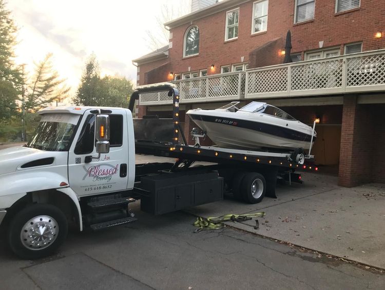 A white tow truck hauling a small boat parked near a multi-story house with a balcony.