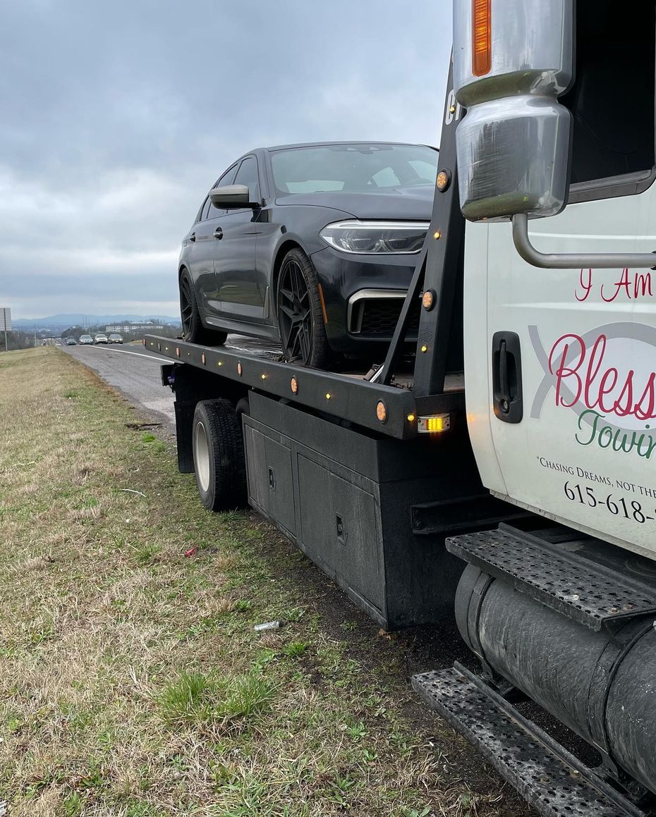 A black car loaded on a tow truck, on a road with grass on the side, under an overcast sky.