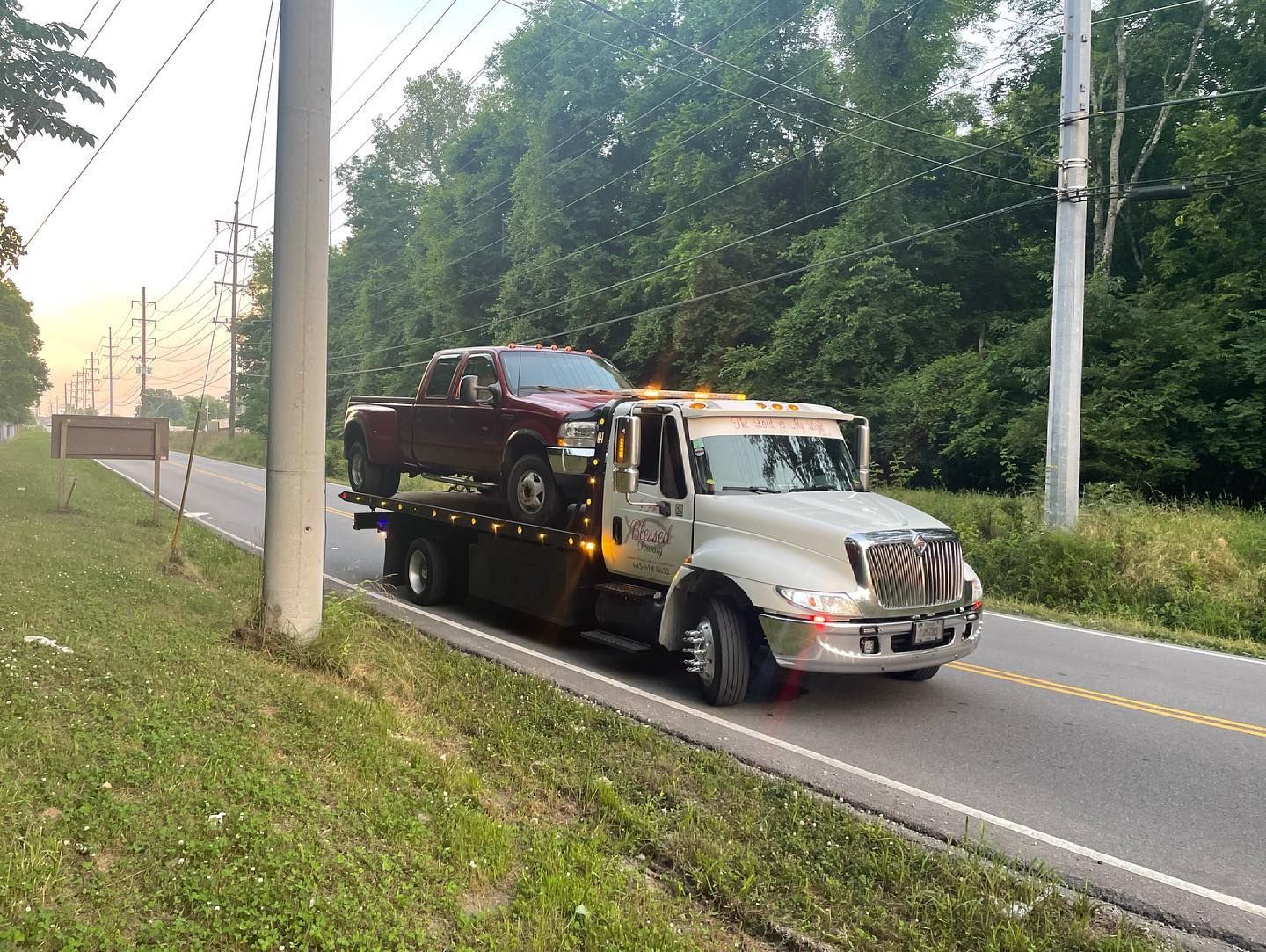 Tow truck with a red pickup truck on its flatbed, driving on a road lined with grass and trees.
