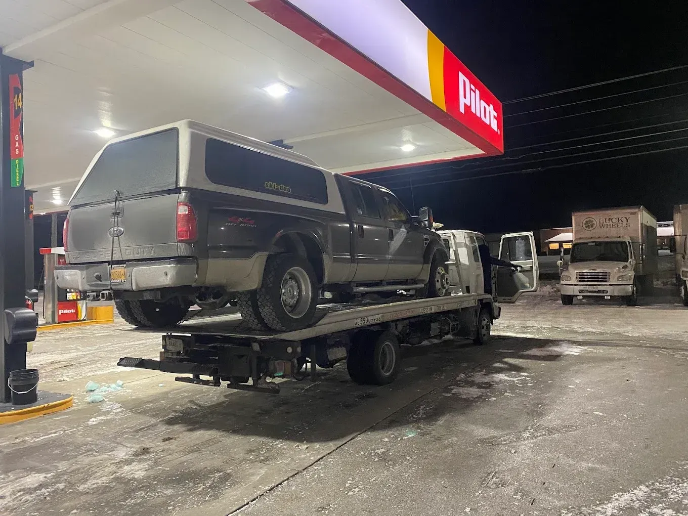 Truck with a camper shell being towed on a flatbed tow truck at a Pilot gas station at night.