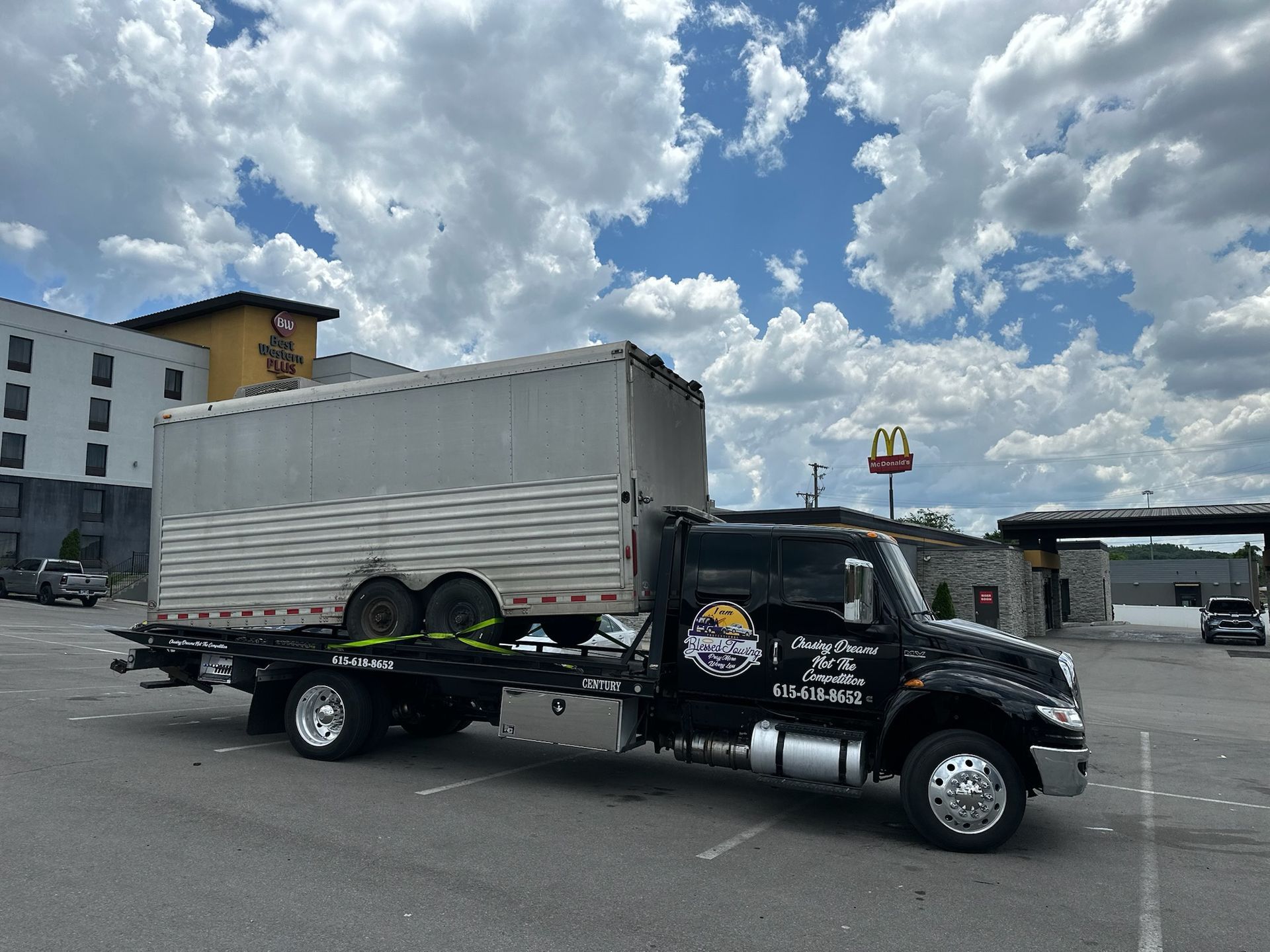 Black tow truck hauling a silver horse trailer in a parking lot on a sunny day.