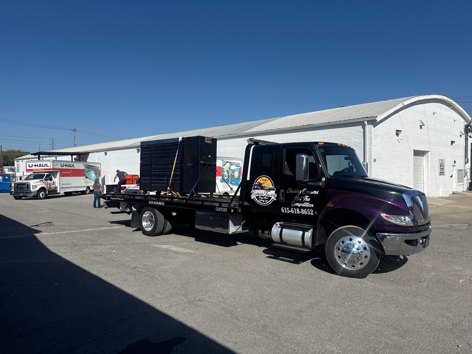 Tow truck with a flatbed carrying large black equipment in front of a white building.