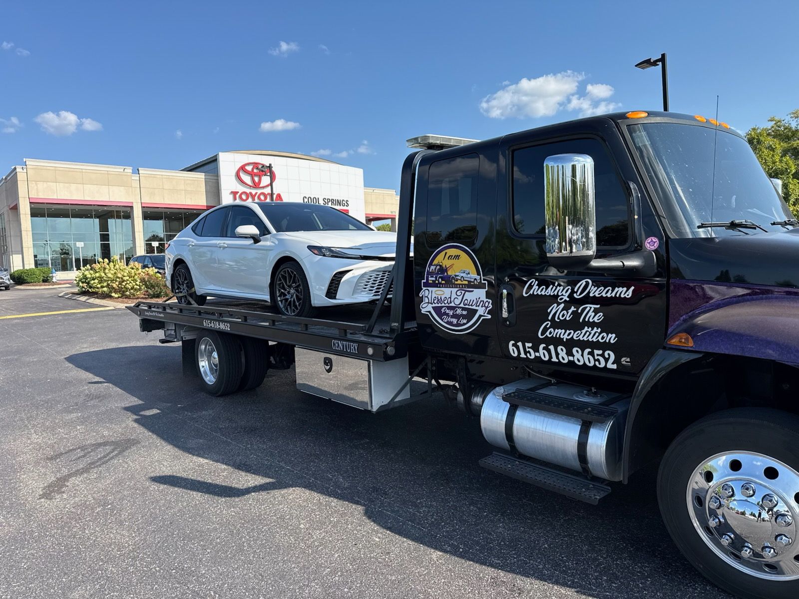 White car on a tow truck in front of a Toyota dealership. Tow truck is black and purple. Sunny day.