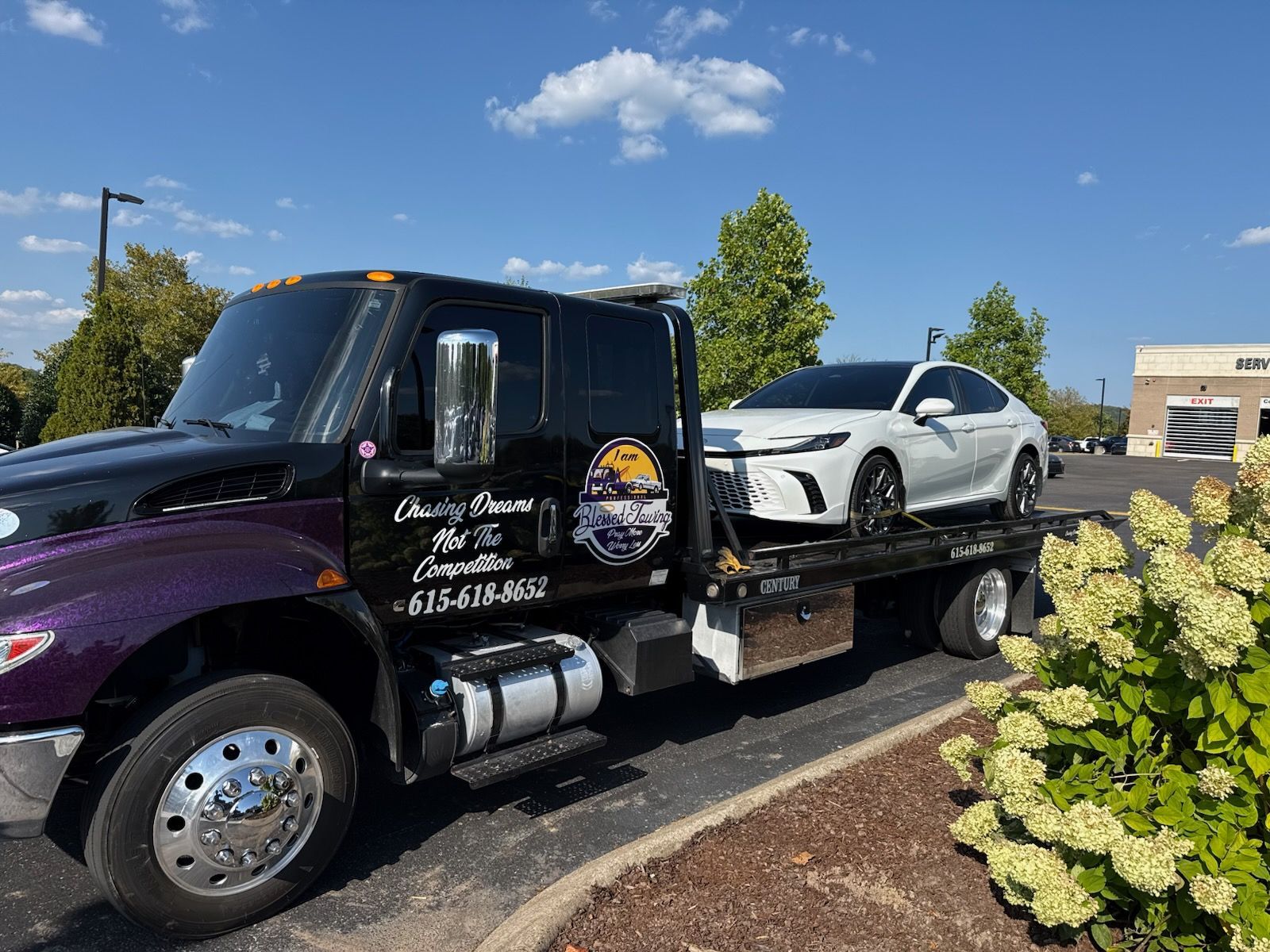 A white car loaded on a black tow truck in a parking lot on a sunny day.
