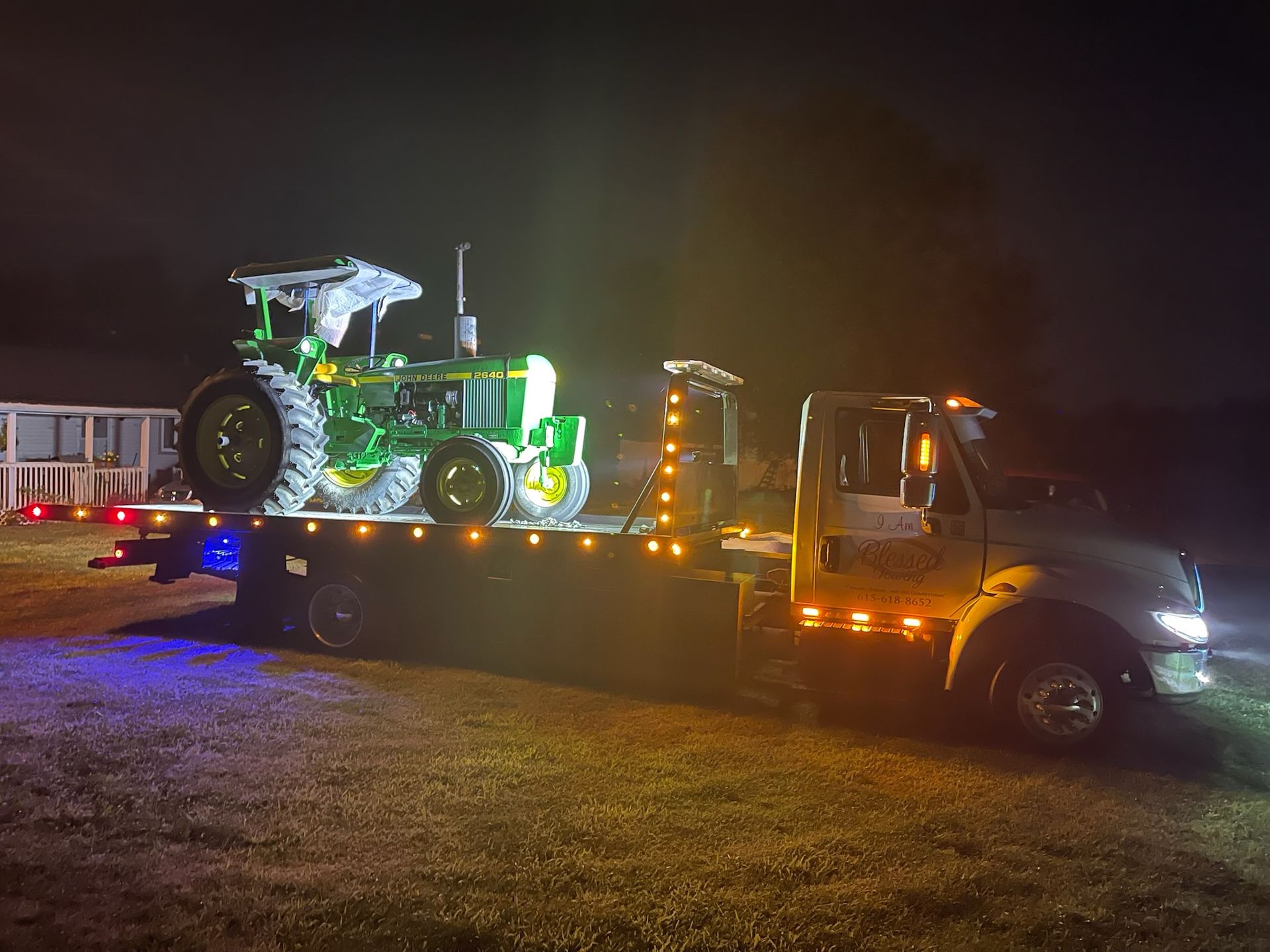 Green and yellow tractor on a flatbed tow truck at night, illuminated by lights.