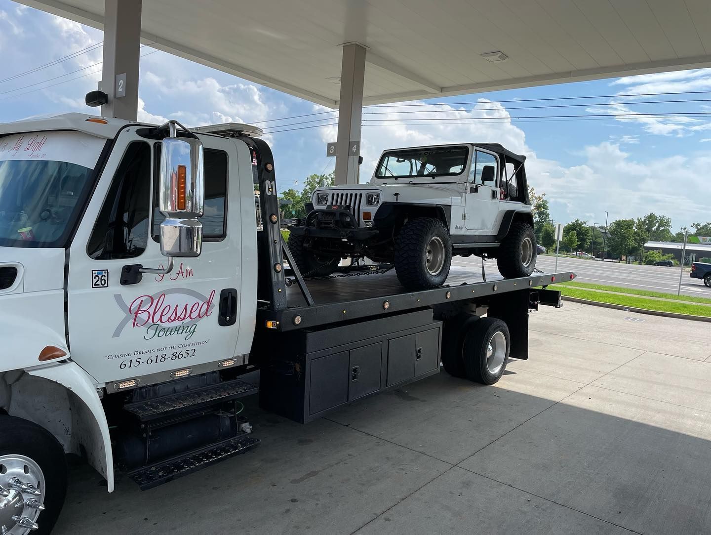 White tow truck with a white Jeep on its flatbed at a gas station.