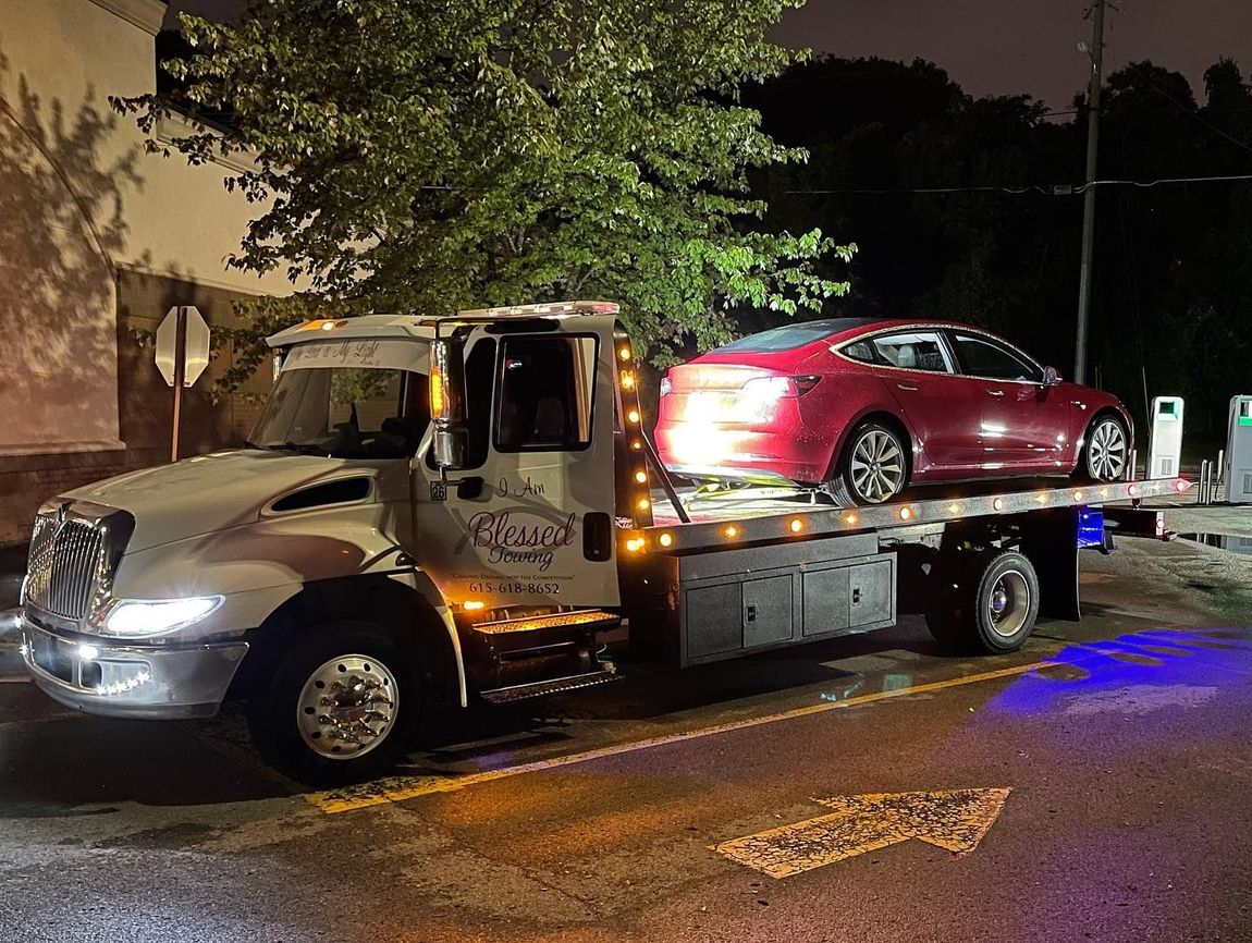 Red Tesla on a tow truck at night. The truck is white with amber lights.
