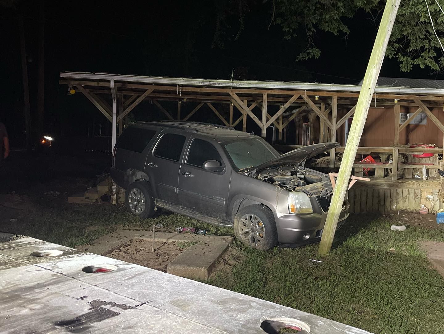 Gray SUV crashed into a wooden structure at night; hood open, damage visible.