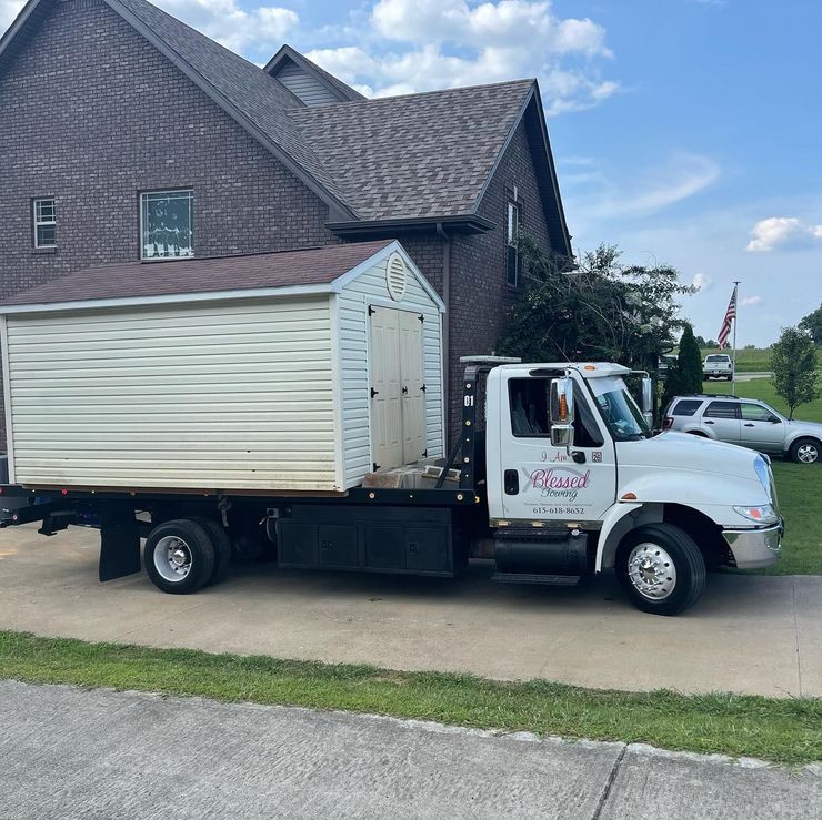 White truck hauling a shed in front of a house on a sunny day.