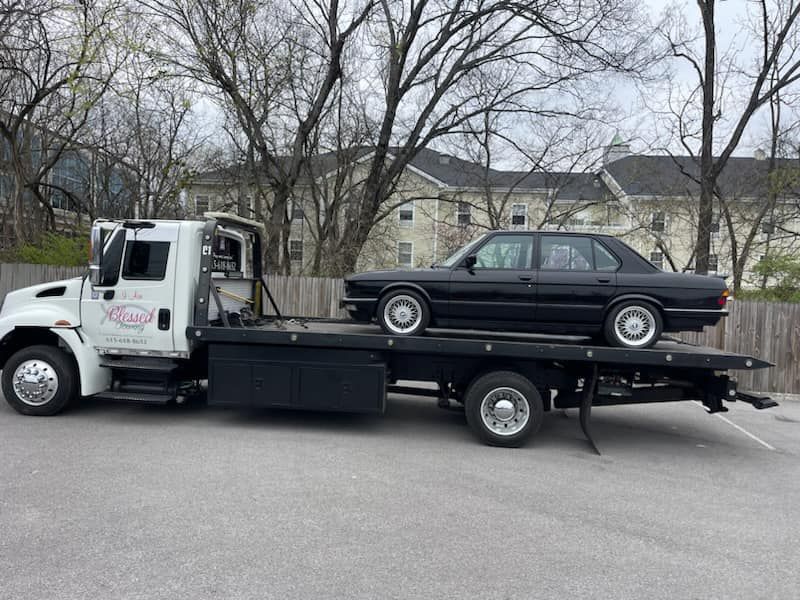 Black sedan on a flatbed tow truck. The truck is white, parked outdoors in front of trees and a building.