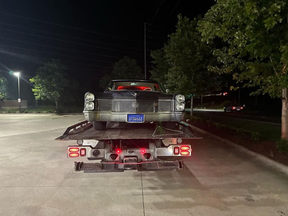 A black classic car on a tow truck at night. Tail lights on, near trees and a street.